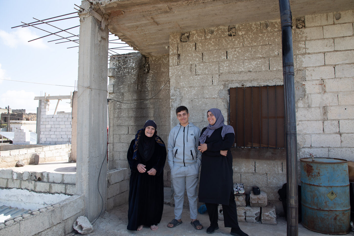 Three members of a family — an older woman, a teenage boy and a woman in a headscarf — stand together outside their unfinished breeze-block house.