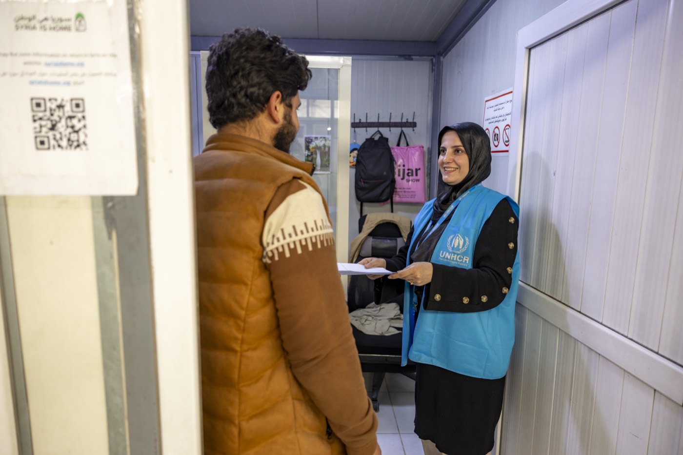 A UNHCR staff member in a blue vest hands a printed form to a man at the counter of a reception and registration office.