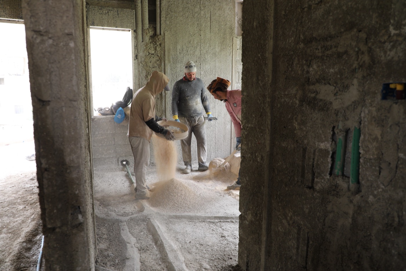 Three construction workers sift and shovel sand inside an unfinished concrete building, a UNHCR-branded bag resting on a ledge behind them.