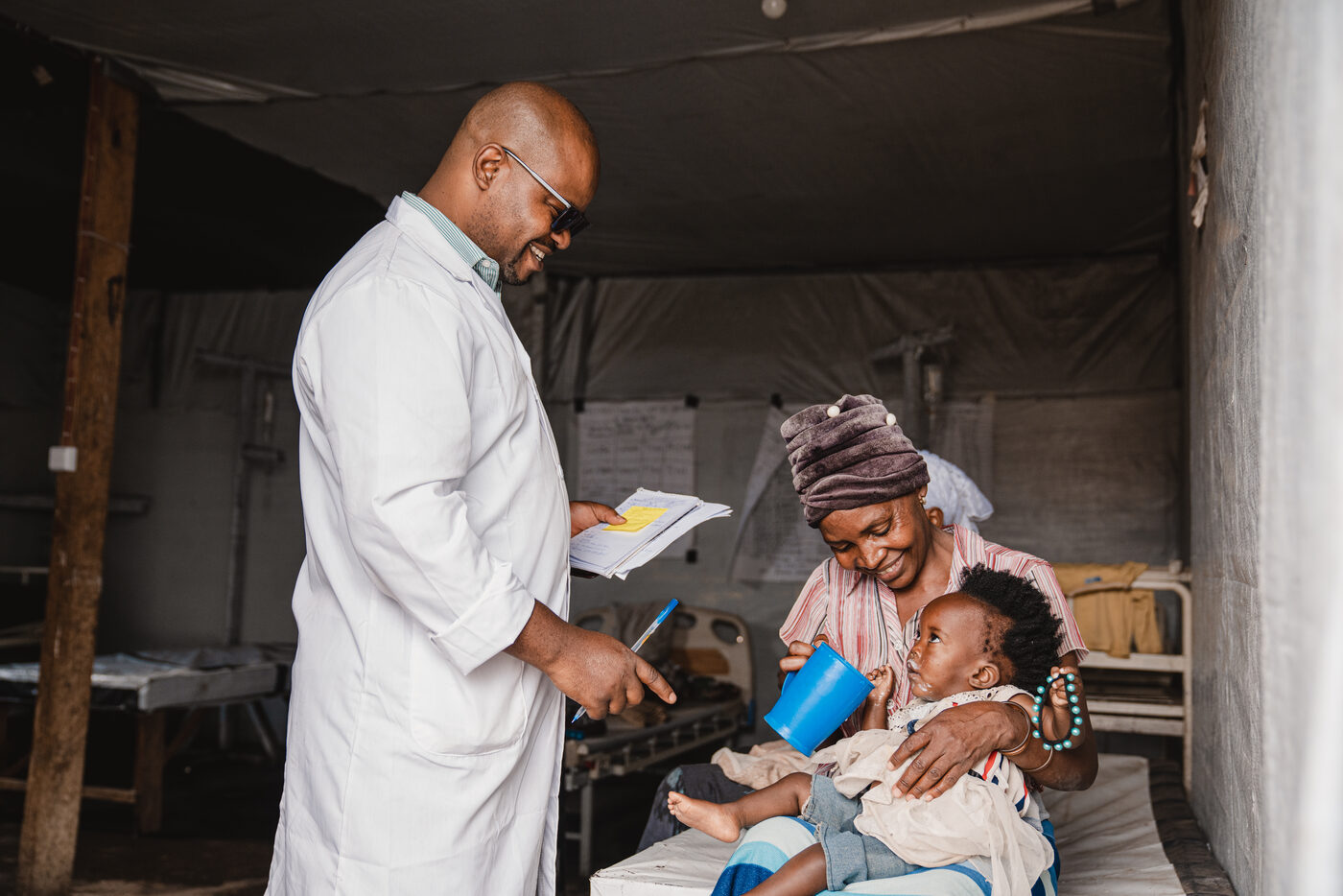 A health worker in a white coat reads from a patient file while a mother, holding a cup for her small child, sits with her on a bed in a field clinic.