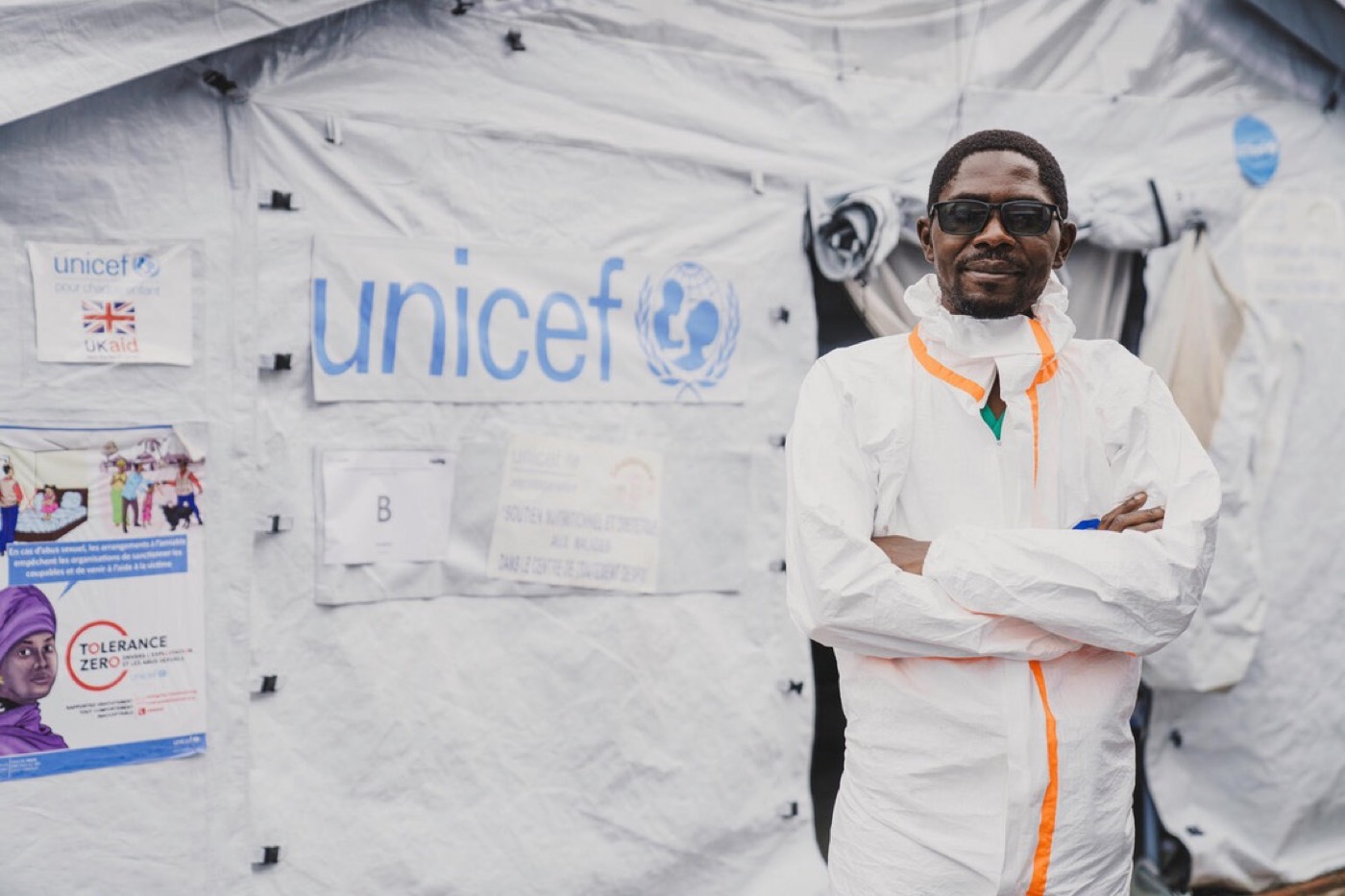 A UNICEF field worker in protective coveralls stands with arms folded in front of a UNICEF-branded tent hung with public health posters.