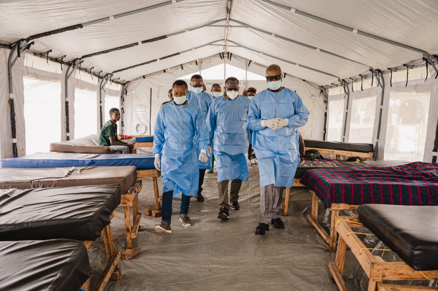 Health workers in gowns and masks walk together down the central aisle of a field treatment tent lined with empty beds.