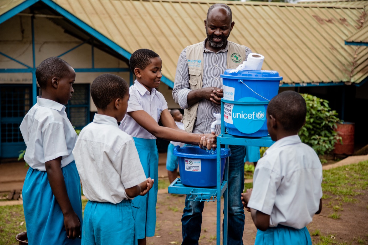 A field officer helps a group of schoolchildren in uniform wash their hands at a UNICEF handwashing station in a school yard.
